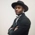 Portrait of african american man standing with his arms crossed in black jacket and hat. Young male model posing on gray background. Vertical studio photography from a DSLR camera. Sharp focus on eyes.